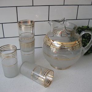 May include: A set of four frosted glass pitcher and tumblers with gold trim. The pitcher has a wide mouth and a handle. The tumblers are tall and slender.
