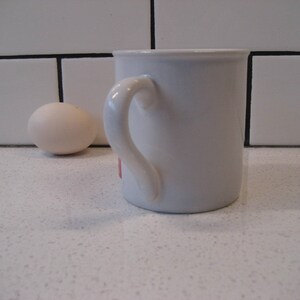 May include: A white ceramic mug with a rounded handle. The mug is sitting on a white countertop.