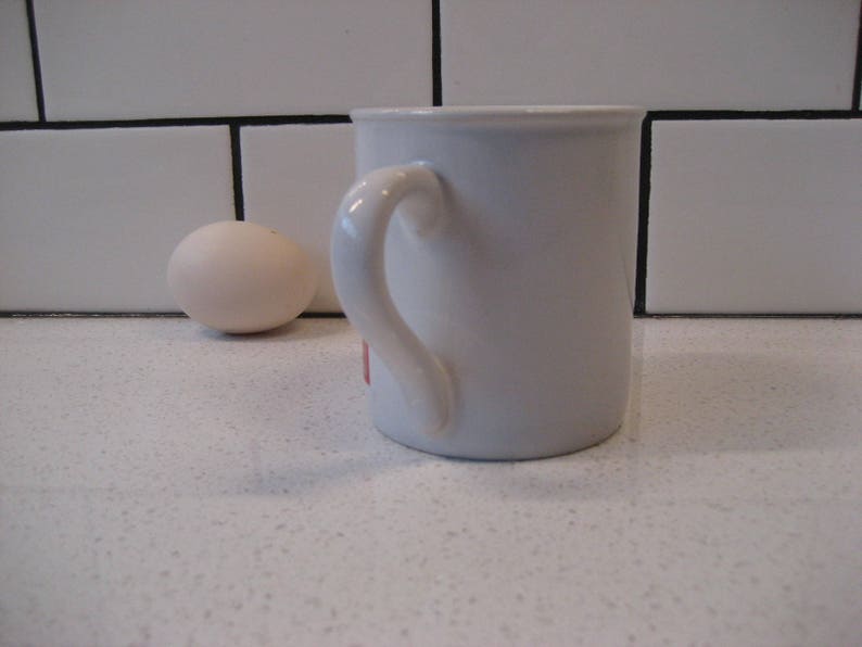May include: A white ceramic mug with a rounded handle. The mug is sitting on a white countertop.