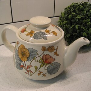 May include: A white ceramic teapot with a floral design featuring yellow, orange, and blue flowers. The teapot has a lid and a spout.