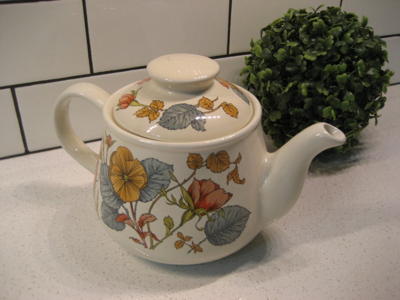 May include: A white ceramic teapot with a floral design featuring yellow, orange, and blue flowers. The teapot has a lid and a spout.