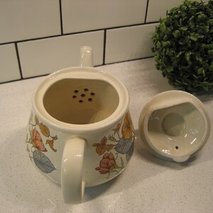 May include: A white ceramic teapot with a floral design and a strainer. The teapot has a lid with a small hole in the center. The teapot is sitting on a white countertop.