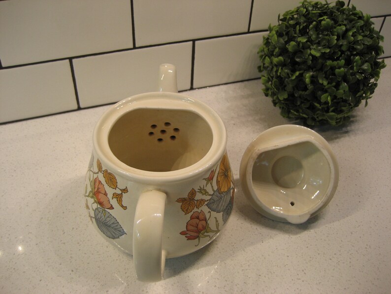 May include: A white ceramic teapot with a floral design and a strainer. The teapot has a lid with a small hole in the center. The teapot is sitting on a white countertop.