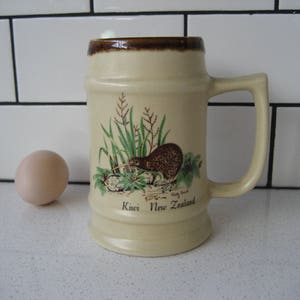 Könnte beinhalten: Ein beigefarbener Bierkrug aus Keramik mit braunem Rand und Henkel. Der Krug zeigt eine grüne und braune Illustration eines Kivis in einem grasbewachsenen Feld. Der Text "Kiwi Neuseeland" ist auf der Vorderseite des Kruges aufgedruckt.
