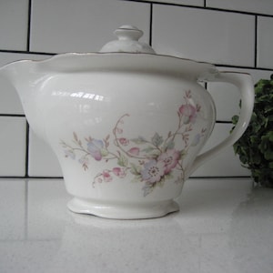 Pode incluir: Uma chaleira branca com um design floral. A chaleira tem uma tampa e uma alça. A chaleira está em cima de uma bancada branca.