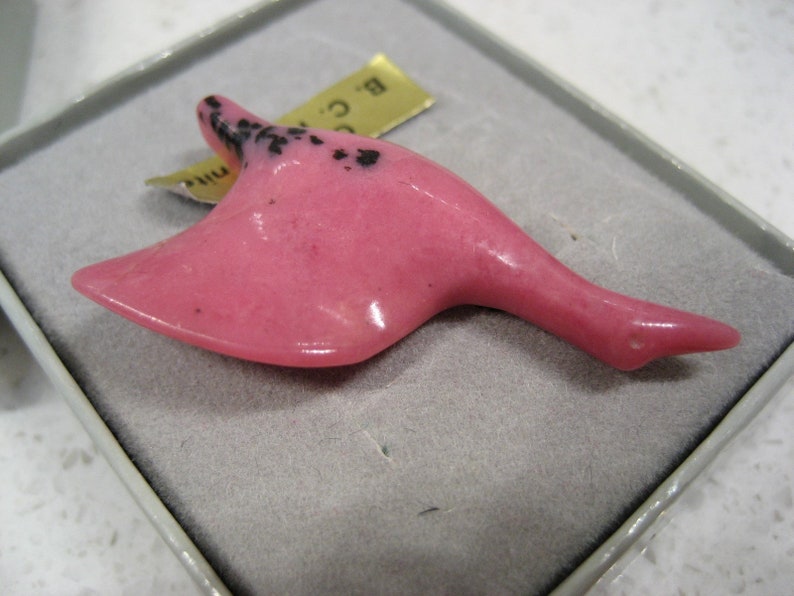 May include: A pink carved goose figurine with black speckles. The figurine is in a small white box with a gold label that reads "B.C. Mini".