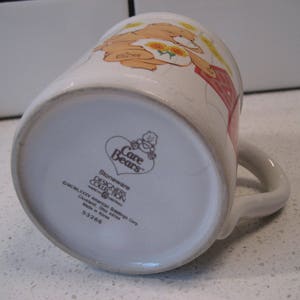May include: White ceramic mug with a Care Bears design featuring a bear with a flower crown and a red dress. The mug has a brown handle and the text "Care Bears Stoneware Designers Collection" on the bottom.