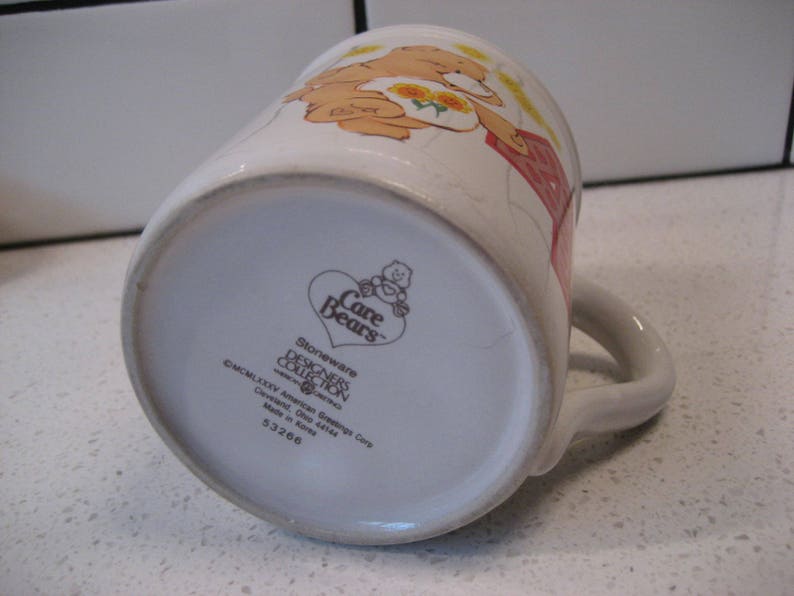 May include: White ceramic mug with a Care Bears design featuring a bear with a flower crown and a red dress. The mug has a brown handle and the text "Care Bears Stoneware Designers Collection" on the bottom.