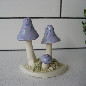 Könnte beinhalten: Ein Set aus drei Keramikpilzen mit violetten und weißen, gefleckten Hüten. Die Pilze sind auf einem weißen Sockel mit grünen Grasakzenten angeordnet.