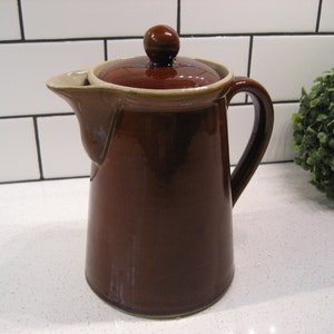 May include: A brown ceramic coffee pot with a lid and a handle. The pot has a light brown rim and a slightly darker brown body.