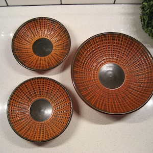 Puede incluir: Tres cuencos de cerámica con un patrón tejido en tonos marrones y naranjas. Los cuencos están anidados, con el más grande en la parte inferior y el más pequeño en la parte superior.