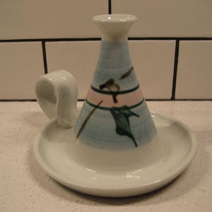 Peut inclure: Un bougeoir en céramique blanche avec un motif floral bleu et rose et une poignée. Le bougeoir a une base large et une ouverture étroite pour une bougie.