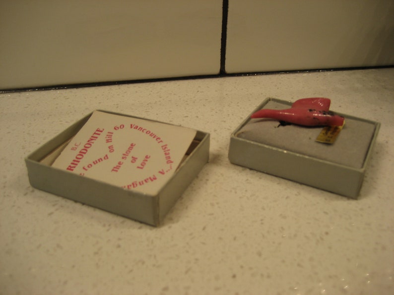 May include: A small pink stone, labeled "Rhodonite", found on "the stone of love" on Galiano Island, BC. The stone is in a small, white box with a label that reads "60 Vancouver Island".