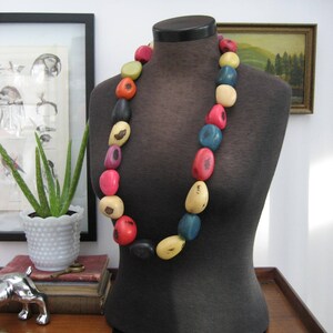 Puede incluir: Un collar colorido con cuentas grandes de forma irregular en varios colores, incluyendo rojo, amarillo, azul y negro, exhibido en un maniquí negro. Las cuentas parecen ser de madera o un material similar.