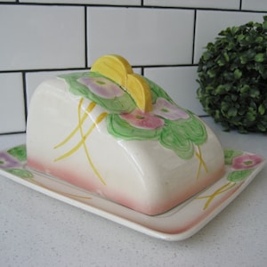 May include: A vintage ceramic butter dish with a domed lid. The lid features a yellow and green floral design with pink flowers. The dish has a matching base with a pink and yellow floral design.