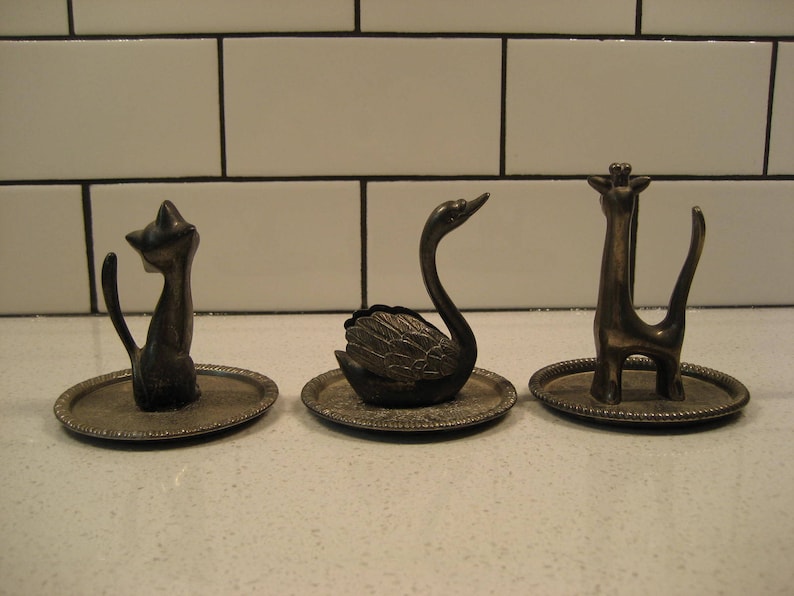 K&ouml;nnte beinhalten: Drei silberne Ringhalter, jeder mit einem anderen Tier: eine Katze, ein Schwan und eine Giraffe. Die Halter stehen auf kleinen, runden, silbernen Tabletts.