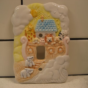 Könnte beinhalten: Ein Keramikschalter mit einer bunten Darstellung der Arche Noah mit Tieren, darunter eine Giraffe, Löwen und ein Zebra. Die Arche ist von Wolken und einer Sonne umgeben.