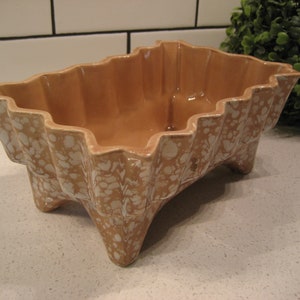 Puede incluir: Un macetero de cerámica marrón con un esmalte blanco moteado. El macetero tiene un borde festoneado y tres patas.