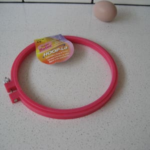 May include: Pink plastic embroidery hoop with a label that reads "Super Busters Hoop-La Embroidery Hoop & Frame.  Cerceaux & cadres en bois.  Hoops y aros para bordar."