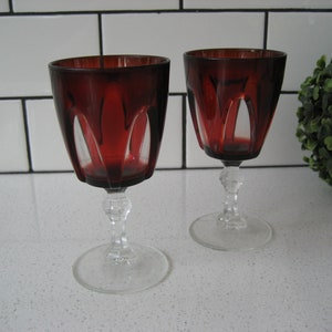 Op de afbeelding: Twee heldere glazen wijnglazen met een diepe rode tint en een geslepen glasontwerp. De glazen hebben een steel.