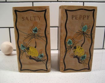 May include: Two wooden salt and pepper shakers with black trim and painted pine tree designs. The shakers are labeled "Salty" and "Peppy".