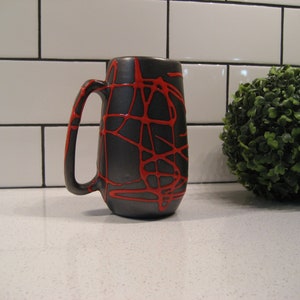K&ouml;nnte beinhalten: Eine schwarze Keramiktasse mit einem roten abstrakten Design. Die Tasse hat einen abgerundeten Griff und einen breiten Boden.