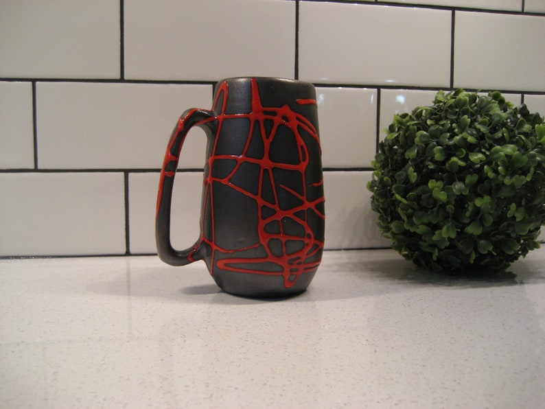 K&ouml;nnte beinhalten: Eine schwarze Keramiktasse mit einem roten abstrakten Design. Die Tasse hat einen abgerundeten Griff und einen breiten Boden.
