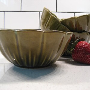 May include: Four green ceramic bowls with a fluted design, stacked on top of each other. Two red strawberries are in the foreground.
