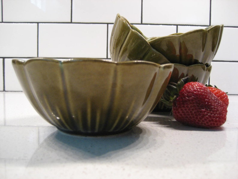 May include: Four green ceramic bowls with a fluted design, stacked on top of each other. Two red strawberries are in the foreground.