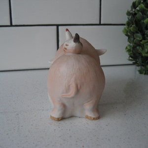 May include: A ceramic pig figurine in a light pink hue, viewed from the rear. The pig has small ears, a curled tail, and a small piglet riding on its back. The figurine has a glossy finish and is set against a white background.