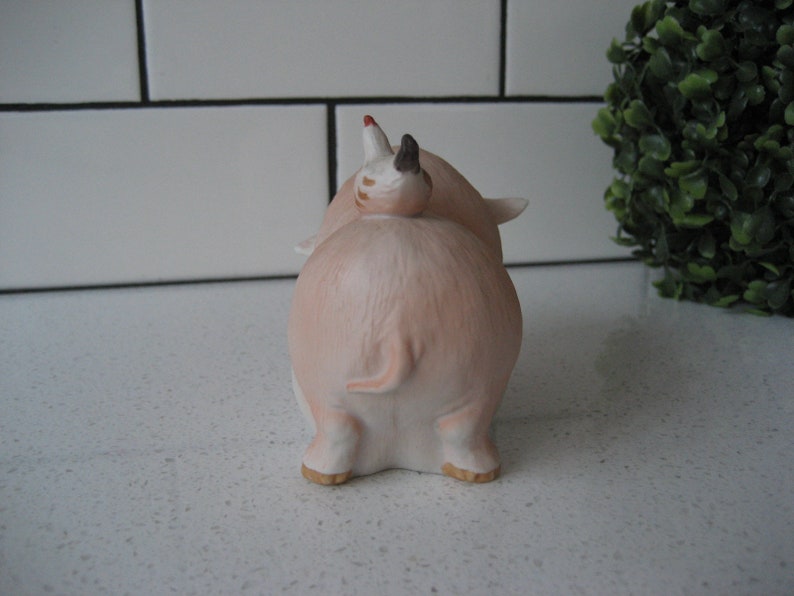 May include: A ceramic pig figurine in a light pink hue, viewed from the rear. The pig has small ears, a curled tail, and a small piglet riding on its back. The figurine has a glossy finish and is set against a white background.