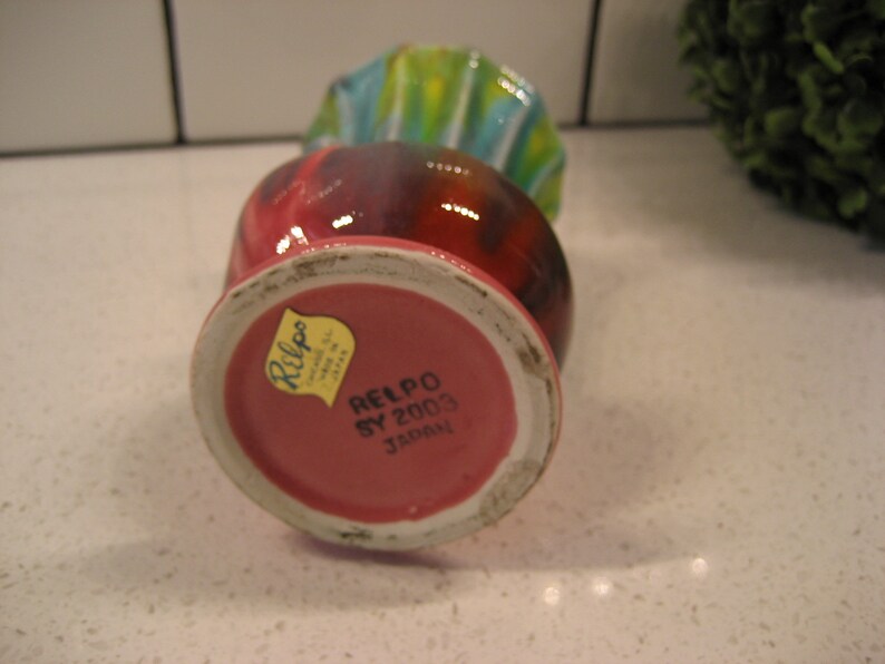 May include: A pink and red ceramic vase with a yellow label that reads "Relpo SY 2003 Japan". The vase has a colourful, abstract design with blue, green, and yellow stripes.