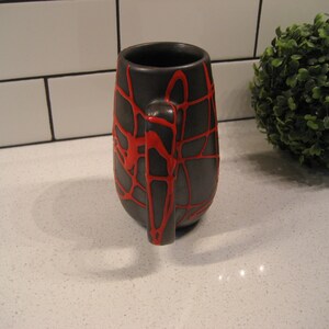 K&ouml;nnte beinhalten: Eine schwarze Keramiktasse mit einem roten Spinnennetz-Design. Die Tasse hat eine abgerundete Form und einen Henkel.