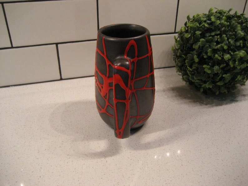K&ouml;nnte beinhalten: Eine schwarze Keramiktasse mit einem roten Spinnennetz-Design. Die Tasse hat eine abgerundete Form und einen Henkel.