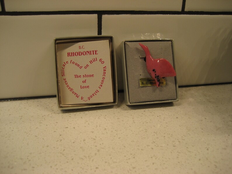 May include: A pink rhodonite bird figurine in a small box. The box has a label that reads "B.C. Rhodonite found on Hill 60 Vancouver Island. Manganese Silicate. The stone of Love."