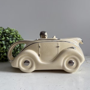 Puede incluir: Tetera de cerámica color crema con forma de coche antiguo. La tetera presenta una figura de conductor, ruedas plateadas y ribetes negros. En el fondo se ve una planta verde y frondosa.