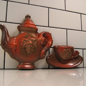 May include: A red and brown ceramic teapot and cup set with a grapevine design. The teapot has a spout and handle, and the cup has a handle. The set is on a white tiled background.