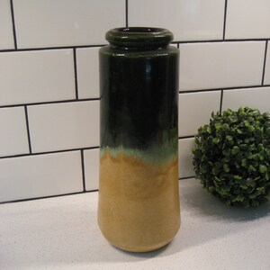 May include: A ceramic vase with a green and brown glaze. The vase is tall and slender with a rounded bottom. It is sitting on a white countertop.