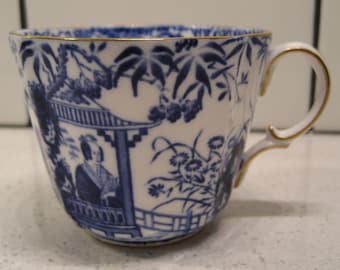 May include: A white teacup with a blue and gold trim. The cup features a Japanese-inspired design with a woman in a blue kimono standing in a pagoda.