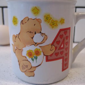 May include: A white ceramic mug with a cartoon bear holding flowers and the number 4. The bear is brown with a pink nose and yellow stars are around the bear. The number 4 is made of red hearts.
