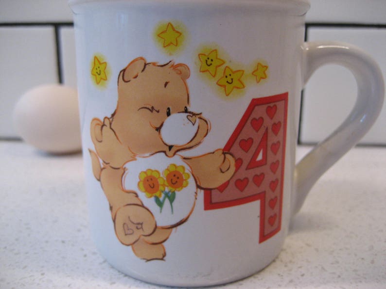 May include: A white ceramic mug with a cartoon bear holding flowers and the number 4. The bear is brown with a pink nose and yellow stars are around the bear. The number 4 is made of red hearts.