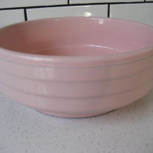 Op de afbeelding: Een roze keramische kom met een witte rand en witte strepen. De kom is leeg en staat op een wit betegeld oppervlak.