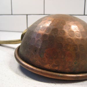May include: A hammered copper saucepan with a brass handle. The saucepan has a rounded bottom and a flared rim.
