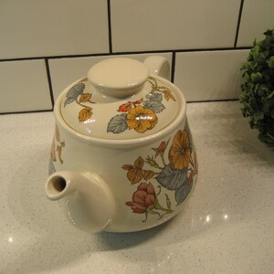 May include: A white ceramic teapot with a floral design featuring yellow, orange, and pink flowers on a green and blue background. The teapot has a lid and a spout.