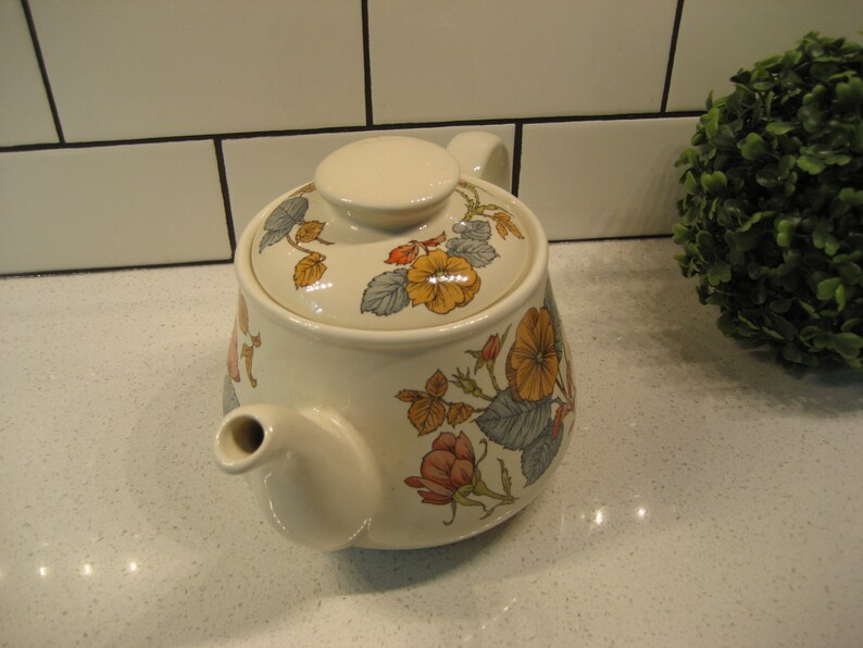 May include: A white ceramic teapot with a floral design featuring yellow, orange, and pink flowers on a green and blue background. The teapot has a lid and a spout.