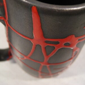 K&ouml;nnte beinhalten: Eine schwarze Keramiktasse mit einem roten Spinnennetz-Design. Die Tasse hat einen breiten Rand und einen Henkel.