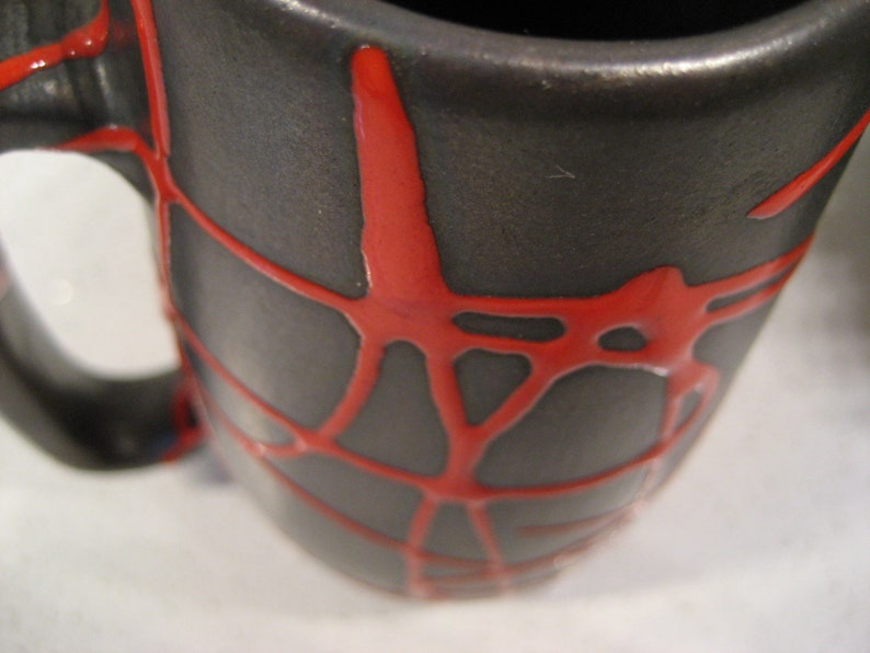 K&ouml;nnte beinhalten: Eine schwarze Keramiktasse mit einem roten Spinnennetz-Design. Die Tasse hat einen breiten Rand und einen Henkel.