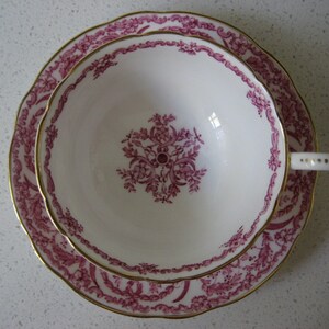 May include: A pink and gold teacup and saucer set with a floral pattern. The cup and saucer have a delicate, scalloped edge.