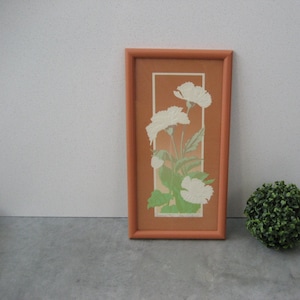 May include: A framed print of white flowers with green leaves on a brown background. The frame is a light orange color.