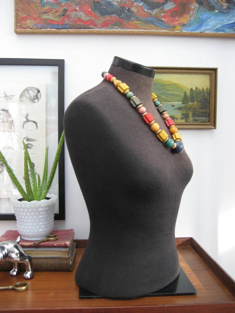 May include: A black fabric mannequin torso with a colorful wooden bead necklace. The necklace has red, yellow, green, blue, and pink beads.